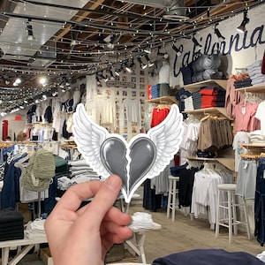 May include: A sticker featuring a gray, broken heart with white wings. The sticker is held in front of a clothing store with racks of shirts, pants, and other apparel. The store has the words "California" on the wall.