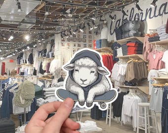 May include: A sticker featuring a sheep wearing a gray hoodie and sitting on a skateboard. The sheep has a small star tattoo. The background shows a clothing store with racks of shirts and jeans.