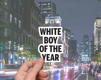 May include: A white sticker with the text "WHITE BOY OF THE YEAR" in a bold, black font. The sticker is held in front of a city street scene at night, with buildings and streetlights visible.
