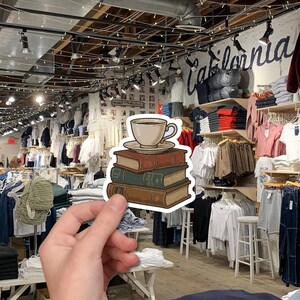 May include: A sticker featuring a stack of three books with a teacup and saucer on top. The books are brown, green, and red, with the teacup and saucer in a light beige color. The sticker is held in front of a clothing store.