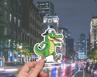 May include: A green alligator cartoon sticker wearing a backwards baseball cap and red sneakers, riding a skateboard. The alligator has a yellow belly and a red tongue sticking out. The sticker is held in front of a city street at night.