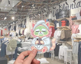 May include: A sticker featuring a cartoon cat wearing a spa robe and face mask, holding a smartphone. The cat has pink hair and paws, with a green face mask. The background shows a clothing store.