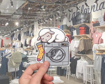 May include: A sticker featuring a cartoon sheep sleeping on a washing machine, with a yellow sock with pink stars. The background shows a clothing store with racks of shirts and jeans.