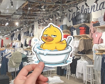 May include: A sticker featuring a yellow cartoon duck relaxing in a white bathtub with bubbles. The duck has a slightly grumpy expression. The sticker is held in front of a clothing store.