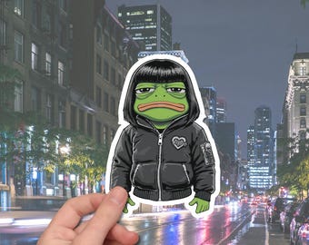 May include: A die-cut sticker featuring a green frog character with black hair and a black jacket. The frog has a neutral expression. The jacket has a heart-shaped patch. The background shows a city street at night.