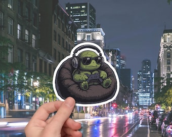 May include: A sticker featuring a cartoon turtle wearing sunglasses and headphones, holding a game controller while seated in a beanbag chair. The sticker is held in front of a city street scene at night.