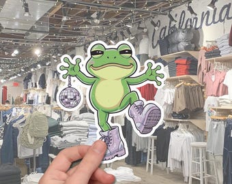 May include: A sticker featuring a cartoon frog with a green body, light green belly, and purple boots. The frog is holding a disco ball and has a relaxed expression. The background shows a clothing store with racks of apparel.