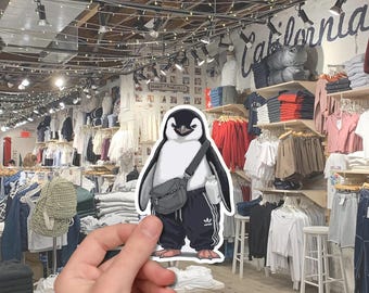 May include: A sticker of a penguin wearing a black crossbody bag, black sweatpants with white stripes, and a white bottle. The penguin has a black and white body and a stern expression. The background is a clothing store.