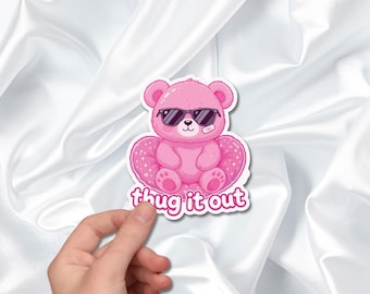 May include: A pink sticker featuring a cartoon teddy bear wearing sunglasses and sitting on a heart-shaped pillow. The text "thug it out" is below the bear. The sticker is held by a hand against a white, silky background.