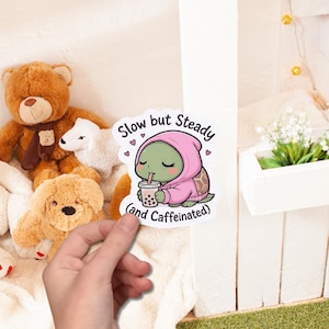 May include: A sticker featuring a cartoon turtle wearing a pink hoodie and drinking boba tea. The sticker has the text "Slow but Steady (and Caffeinated)" with heart accents. The sticker is held by a hand.