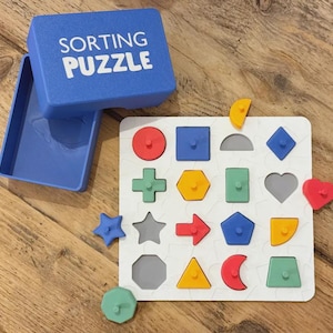 May include: A blue sorting puzzle with a matching box. The puzzle board is white with various colored geometric shapes, including circles, squares, and stars. Some shapes are placed on the board, while others are loose.