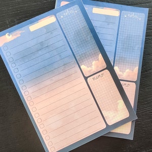 May include: Two blue and pink notepads with a moon and clouds design. The notepads have sections for notes and memos. The notepads have a grid pattern.