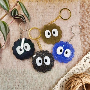Peut inclure: Quatre porte-clés représentant un personnage rond, flou et de dessin animé avec de grands yeux. Les porte-clés sont en noir, gris foncé, or et bleu.