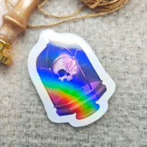 Könnte beinhalten: Ein holografischer Aufkleber mit einem Schädel mit einem Kristall in einer Glaskuppel. Die Kuppel ist schillernd und hat einen Regenbogeneffekt.