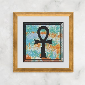 May include: Framed cross-stitch art featuring a black ankh symbol. The ankh is centered on a colorful background with blue, orange, and white hues. The artwork is framed in a gold-colored, ornate frame, and is displayed against a white marble background.