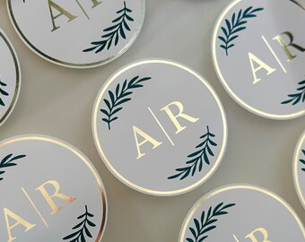 May include: Round white stickers with gold lettering and green leafy designs. The stickers feature the initials "A/R" in a gold script font.
