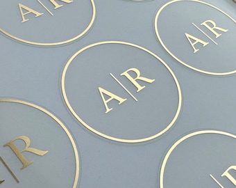 May include: Close-up of a set of round, light gray stickers with a gold outline. Each sticker features the letters "A/R" in gold.