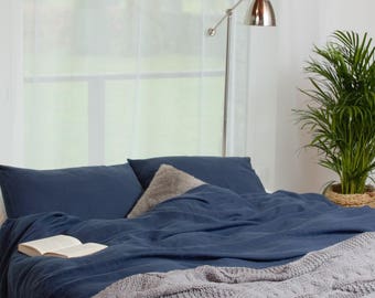 May include: A blue linen duvet cover with a book on top, two blue linen pillowcases, and a gray throw blanket. The bed is in front of a window with white curtains and a floor lamp.