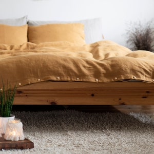 May include: A mustard yellow linen duvet cover with button fastenings on a wooden bed frame. The bed is made with matching pillowcases and a white linen sheet. The bed is in a bedroom with a white rug and a small plant on the floor.