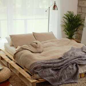 Könnte beinhalten: Ein Bett mit einem hellbraunen Leinen-Bettbezug und einer grauen Strickdecke. Das Bett steht auf einem Holzpalettenrahmen. Auf dem Bett liegen zwei Kissen, eines mit einem weißen Kissenbezug und eines mit einem hellbraunen Kissenbezug. Neben dem Bett steht ein Beistelltisch mit einer Pflanze im Topf.