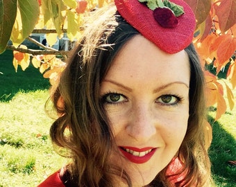 May include: A woman wearing a red dress and a red felt hat with a green leaf and a red berry embellishment. The hat is tilted on her head.