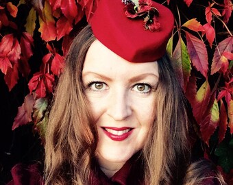 May include: A red pillbox hat with floral accents. The hat is worn by a person with long brown hair and red lipstick. The background features red and yellow autumn leaves.