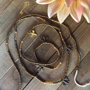 May include: A set of three gold beaded waist beads with bee charms. The beads are arranged on a wooden surface with a sunflower in the background. The text "Treasure Box" is visible in the image.