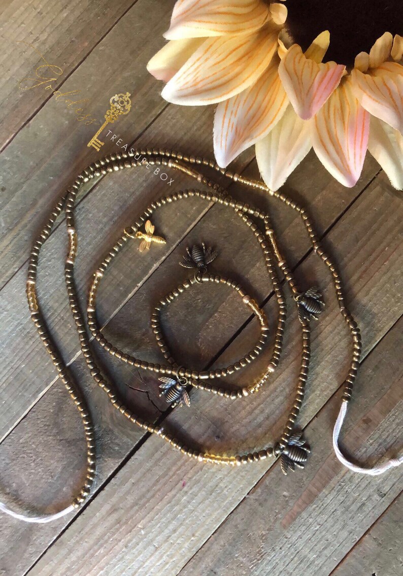 May include: A set of three gold beaded waist beads with bee charms. The beads are arranged on a wooden surface with a sunflower in the background. The text "Treasure Box" is visible in the image.