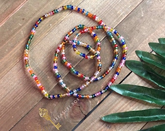Puede incluir: Un collar de cuentas de cintura de colores con múltiples hilos de cuentas en varios colores, incluyendo rojo, naranja, amarillo, verde, azul y morado. Las cuentas están dispuestas en un patrón circular y están enhebradas en un hilo transparente. Las cuentas están sobre una superficie de madera.