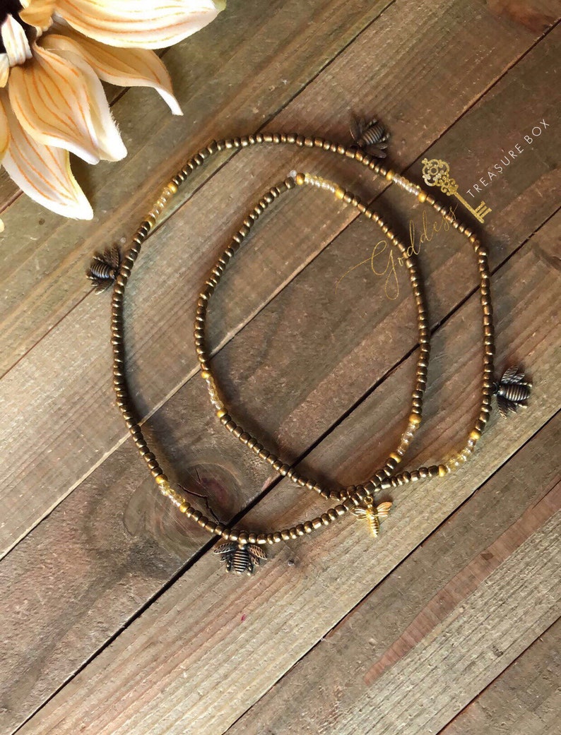 May include: Two gold beaded waist beads with bee charms. The beads are strung on a brown cord and the charms are made of metal.