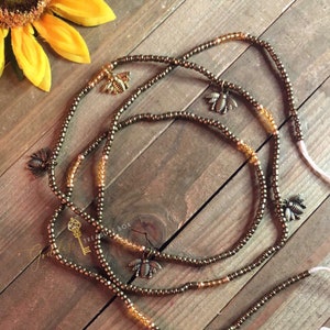 May include: Three brown beaded waist beads with gold bee charms. The beads are strung on a white cord with tassels.