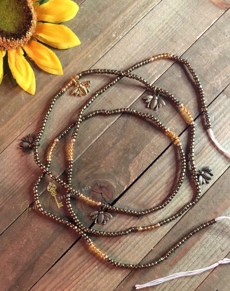 May include: Three brown beaded waist beads with gold bee charms. The beads are strung on a white cord with tassels.