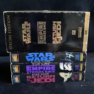 Puede incluir: Una pila de cuatro cintas VHS de la trilogía de Star Wars. Las cintas son negras con letras doradas y muestran los títulos "Star Wars: Una nueva esperanza", "El imperio contraataca" y "El retorno del Jedi".