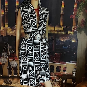 May include: A black and white sleeveless dress with a geometric pattern. The dress has a deep V-neckline and a belt with a silver buckle. The dress is being modeled on a mannequin against a backdrop of a cityscape.