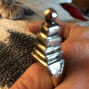 May include: A silver ring with a geometric design that resembles a pyramid. The ring has a rounded top and is worn on a finger.