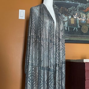 May include: A silver and gray sequined shawl draped over a white mannequin. The shawl features a geometric pattern with vertical and diagonal lines. The fabric has a shimmering effect, and the edges are trimmed with a darker gray material.