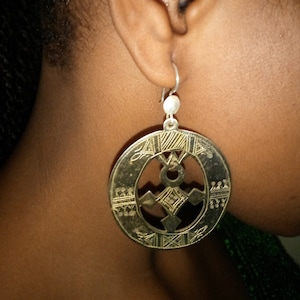 May include: A pair of gold-toned earrings with a circular design featuring a geometric pattern. The earrings are hanging from a silver hook.