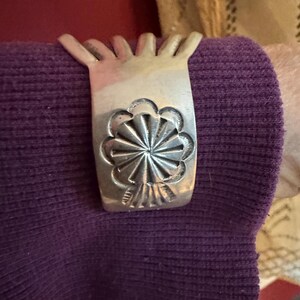 Puede incluir: Un brazalete de plata con un diseño floral decorativo. El brazalete está hecho de múltiples hebras de metal y tiene una forma redondeada.