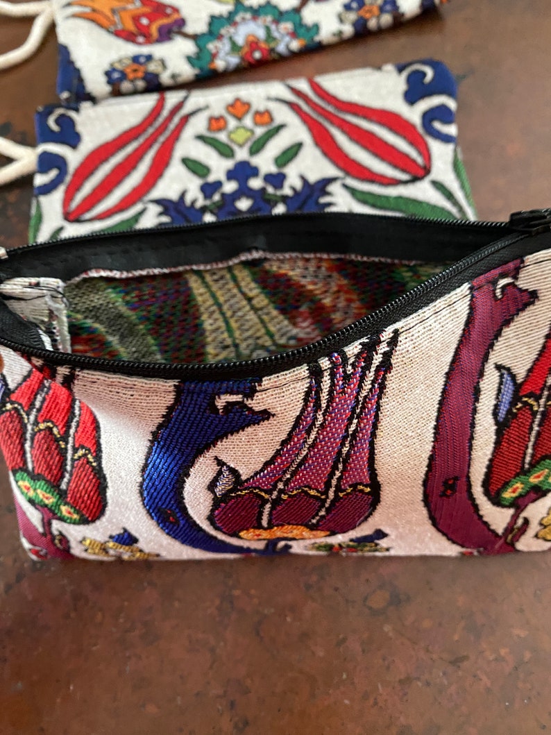 Puede incluir: Una peque&ntilde;a bolsa blanca con cremallera negra. La bolsa est&aacute; decorada con un estampado floral colorido con tulipanes rojos, azules y morados. El estampado recuerda a los textiles turcos tradicionales.