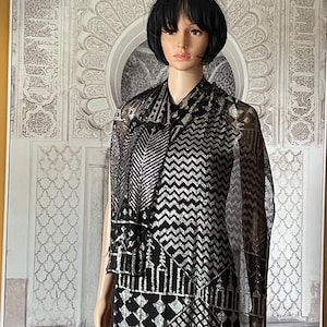 Puede incluir: Un maniquí exhibe un vestido y un chal con diseños en negro y plata. El vestido presenta patrones geométricos y lineales. El chal transparente se extiende sobre los hombros. El fondo muestra una pared decorativa.