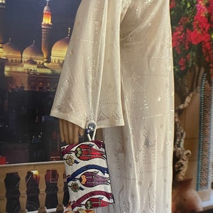 Puede incluir: Un vestido blanco, transparente, de manga larga con un patr&oacute;n geom&eacute;trico. El vestido est&aacute; siendo sostenido por una mano, y un bolso de mano colorido con un dise&ntilde;o floral est&aacute; colgando de la mu&ntilde;eca.