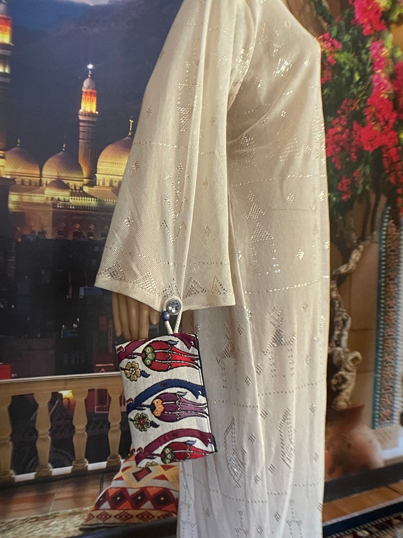 Puede incluir: Un vestido blanco, transparente, de manga larga con un patr&oacute;n geom&eacute;trico. El vestido est&aacute; siendo sostenido por una mano, y un bolso de mano colorido con un dise&ntilde;o floral est&aacute; colgando de la mu&ntilde;eca.