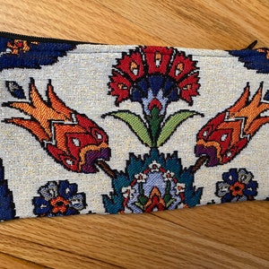 Puede incluir: Una bolsa de tela blanca con cierre de cremallera y un estampado floral colorido. El estampado presenta flores rojas, naranjas, amarillas, azules, verdes y moradas. La bolsa mide aproximadamente 20 cm de largo.