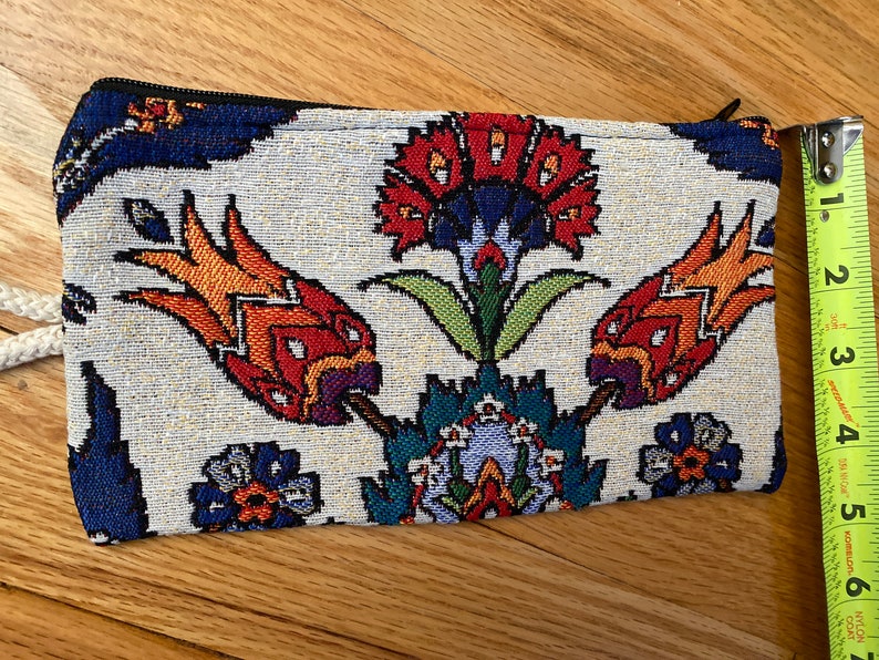 Puede incluir: Una bolsa de tela blanca con cierre de cremallera y un estampado floral colorido. El estampado presenta flores rojas, naranjas, amarillas, azules, verdes y moradas. La bolsa mide aproximadamente 20 cm de largo.