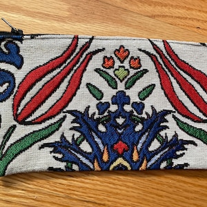 Puede incluir: Una bolsa de tela blanca con cremallera y un dise&ntilde;o floral colorido con flores rojas, azules, verdes y amarillas. La bolsa tiene un cord&oacute;n blanco.