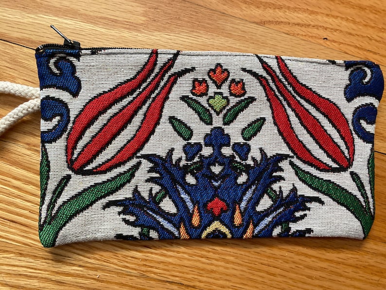 Puede incluir: Una bolsa de tela blanca con cremallera y un dise&ntilde;o floral colorido con flores rojas, azules, verdes y amarillas. La bolsa tiene un cord&oacute;n blanco.