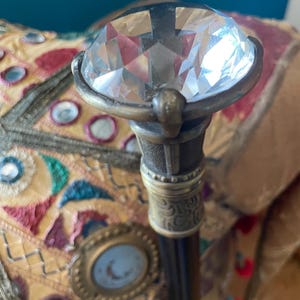 May include: A decorative walking cane with a large, faceted, clear crystal at the top. The handle is dark with ornate metal detailing. The cane rests against a colorful, embroidered surface with jewel-toned accents.