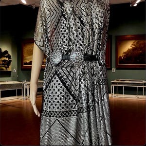 May include: A silver and black patterned dress with a black belt featuring two large, decorative silver buckles. The dress has short sleeves and a geometric design. The dress is displayed on a mannequin.