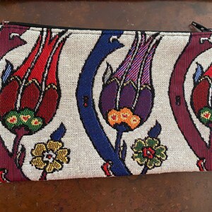 Puede incluir: Una cartera de mano de tela blanca con cierre de cremallera. La cartera presenta un estampado floral colorido con flores rojas, moradas, azules, verdes y amarillas.