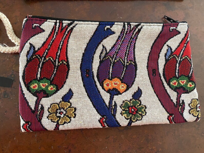Puede incluir: Una cartera de mano de tela blanca con cierre de cremallera. La cartera presenta un estampado floral colorido con flores rojas, moradas, azules, verdes y amarillas.